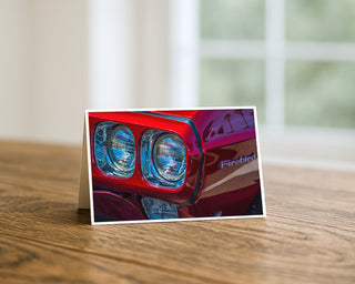 5x7 greeting card featuring close-up photography of a red Pontiac Firebird classic car headlight and emblem with chrome details by Will Davis Studios.