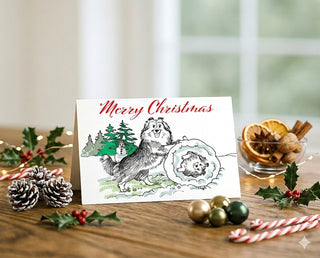 Product mockup of a Will Davis Studios 5x7 Shetland Sheepdog Christmas greeting card standing upright on a warm wooden table surrounded by festive holiday decorations including frosted pine cones, holly sprigs with red berries, gold and green ornament balls, candy canes, fairy lights, and a glass bowl of dried orange slices, cinnamon sticks, and walnuts, featuring a classic pen and ink illustration of two Shelties in a humorous snowball scene with Merry Christmas in red script above, photographed in soft na