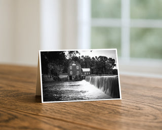 Black and white photo of a house near a waterfall on a wooden surface with a blurred background