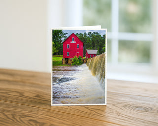 Greeting card with a red house and waterfall on a wooden surface Will Davis Studios