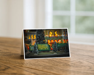 Fine art Christmas greeting card displayed on a wooden table featuring a miniature holiday train with “Merry Christmas” lettering passing through a glowing Christmas village scene. Will Davis Studios