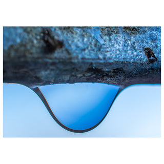 Fine art greeting card featuring an extraordinary macro photograph of a single water droplet suspended in perfect surface tension beneath a rough textured dark stone, its flawless teardrop curve reflecting a luminous pale blue sky just before the moment of release, creating a stunning compositional divide between ancient weathered rock above and pristine pure water below, 5x7 blank card with envelope by Will Davis Studios.