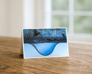 Product mockup of a Will Davis Studios 5x7 fine art greeting card in landscape orientation standing upright on a warm wooden table, featuring a macro photograph of a single water droplet suspended in perfect surface tension beneath a rough dark textured stone against a pristine pale blue background, photographed in soft natural light with a bright window in the background.
